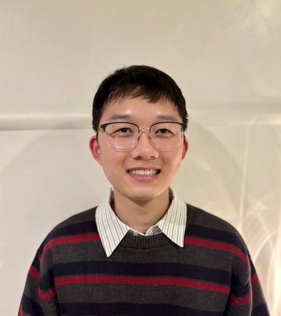 Eunki smiling, wearing glasses and a striped sweater over a collared shirt, against a plain cream wall